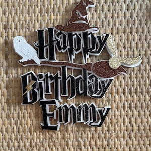 May include: A black and white cake topper with the words "Happy Birthday Emmy" and a Harry Potter theme. The topper features a brown and gold broom, a white owl, and a brown Sorting Hat.
