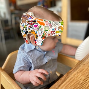 May include: A white infant cranial helmet covered in colorful Nickelodeon cartoon character stickers. The helmet is worn by a baby in a blue striped shirt, seated in a wooden high chair. The helmet has a protective design.