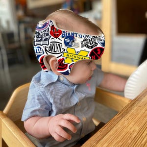 Puede incluir: Un niño pequeño con un casco blanco cubierto de pegatinas de colores, incluyendo logotipos de Nueva York, Brooklyn, Michigan y los Gigantes de Nueva York. El casco está decorado con una variedad de otras pegatinas, incluyendo una cara sonriente, una estrella y una pelota de fútbol. El niño está sentado en una silla alta y mirando hacia la derecha.