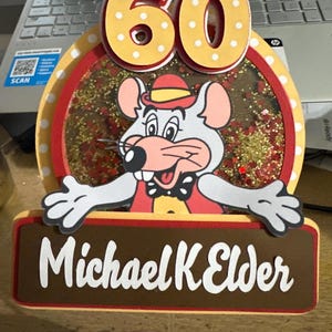 May include: A colorful 60th birthday cake topper featuring a cartoon mouse character. The topper includes the number "60" in yellow with white polka dots, the name "Michael K Elder", and a clear window filled with glitter.