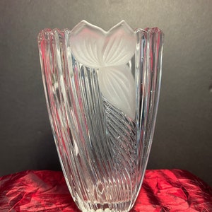 Puede incluir: Jarrón de cristal transparente con un diseño floral esmerilado. El jarrón tiene una forma acampanada con líneas verticales texturizadas. El diseño esmerilado presenta una flor estilizada. El jarrón se exhibe sobre una superficie roja.