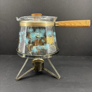 May include: A vintage glass coffee pot with a wooden handle and lid. The clear glass pot features a decorative pattern of blue pine needles and gold pine cones. It sits on a metal stand with a small burner.