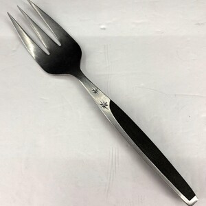 以下が含まれることがあります： 3本歯のステンレス製サービングフォーク。ハンドルには黒い象嵌とスターバーストのデザインが施されています。この食器は、料理を盛り付けるために設計されています。