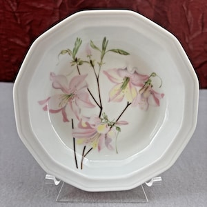 May include: White octagonal plate with a floral design. The plate features a delicate illustration of pink and yellow flowers with green leaves and brown stems. The plate is displayed on a clear stand.