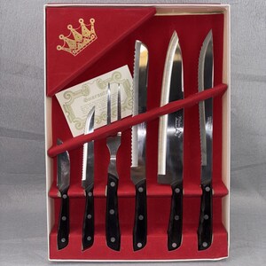 May include: A boxed set of kitchen knives with black handles and stainless steel blades. The set includes various knife types, a fork, and a guarantee card, all presented in a red velvet-lined box with a gold crown emblem.