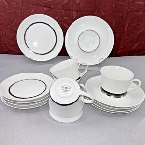Puede incluir: Un juego de té de porcelana blanca con detalles en negro y plata. El juego incluye tazas, platillos y platos. Los platos tienen un borde negro. Las tazas tienen una banda negra alrededor del borde y una banda plateada en la base.