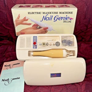 May include: An off-white Electric Manicure Machine kit, "Nail Genie Deluxe", with a variety of attachments. The box features an illustration of a hand using the device. The kit includes a yellow handpiece, accessories, and instruction booklets.