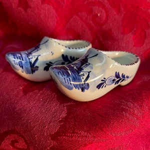 Könnte beinhalten: Ein Paar Miniatur-Holzschuhe, bemalt mit blau-weißen Blumen- und Landschaftsmotiven. Die Holzschuhe sind aus Keramik und werden auf einer roten, gemusterten Oberfläche präsentiert. Die Holzschuhe sind ein traditionelles niederländisches Souvenir.