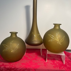 May include: Three glass vases with a frosted green finish and etched leaf designs. The vases are arranged on a red patterned surface.