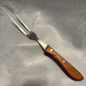 Puede incluir: Un tenedor para trinchar de acero inoxidable con dos púas. El mango es de madera marrón oscuro y está asegurado con tres remaches plateados. El tenedor está diseñado para manipular y servir carnes asadas.