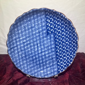 May include: A blue and white decorative plate with a scalloped edge. The plate features two distinct patterns: a diamond grid with starbursts and a geometric floral design. The rim has a brown border.
