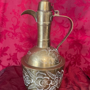 May include: A vintage brass coffee pot with a long spout, curved handle, and a pointed lid. The pot features intricate floral engravings around the base and on the body. The background is a deep red fabric.