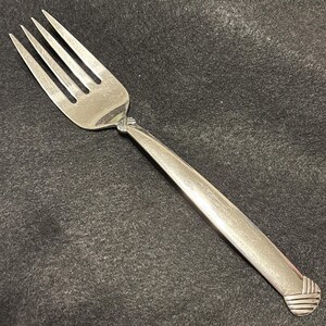 Op de afbeelding: Een zilveren vork met vier tanden en een lange, licht gebogen steel. De steel heeft een textuurontwerp en een decoratief element waar hij de tanden ontmoet. De vork is gemaakt van een glanzend, reflecterend metaal.