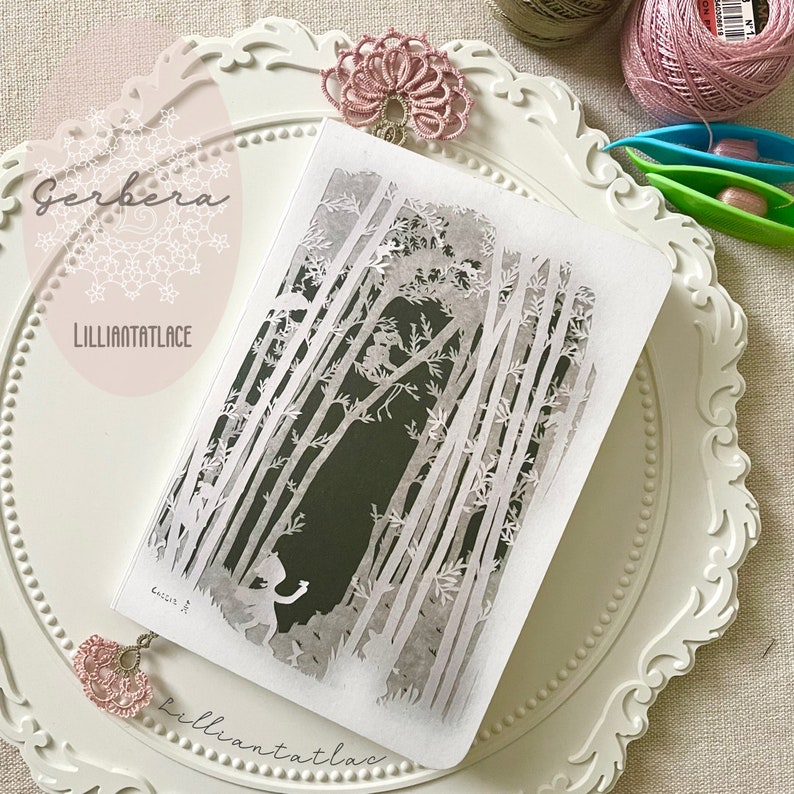 May include: A rectangular notebook featuring a white paper-cut style forest scene with tall trees and a small figure on a dark background. The cover includes the text Gerbera and Lilliantatlace. Two pink lace tatting bookmarks are attached.