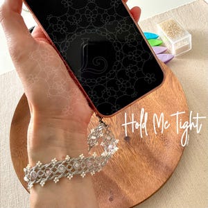 May include: A rose gold phone case with a black screen featuring a floral design and the letter "L". A silver beaded wrist strap is attached to the phone. The wooden surface has the words "Hold Me Tight".