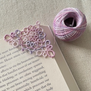 Pu&ograve; includere: Un segnalibro in pizzo delicato, fatto a mano, in un gradiente di rosa e bianco. Il segnalibro ha la forma di un triangolo e presenta un design floreale intricato. Il segnalibro &egrave; appoggiato su un libro.
