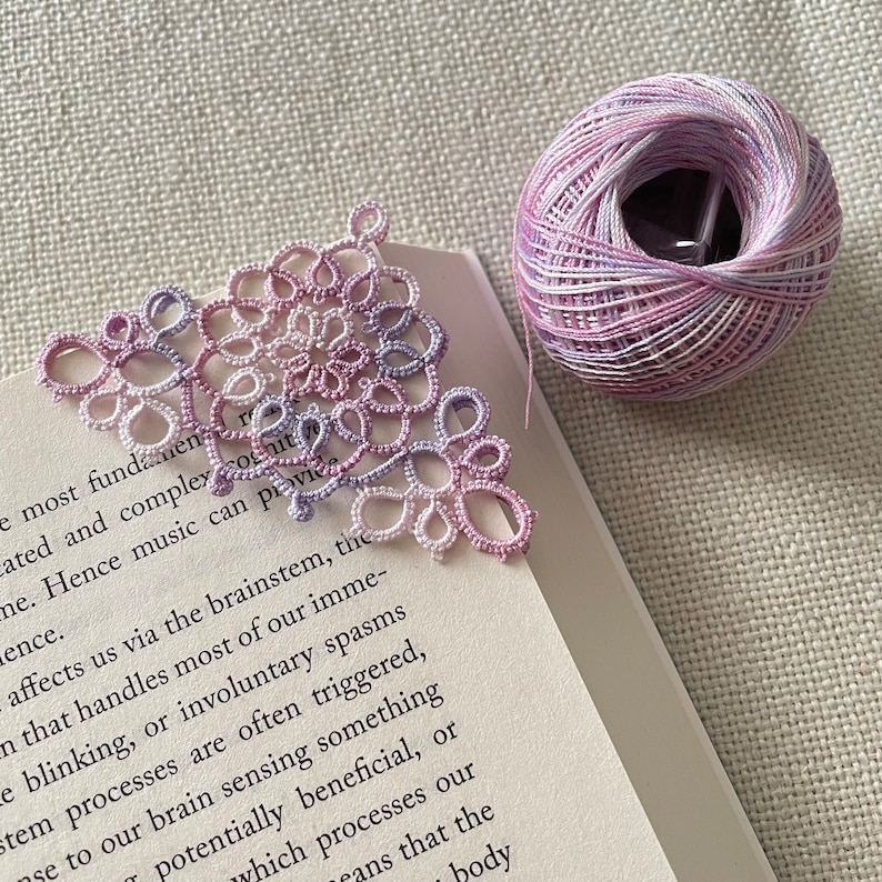 Pu&ograve; includere: Un segnalibro in pizzo delicato, fatto a mano, in un gradiente di rosa e bianco. Il segnalibro ha la forma di un triangolo e presenta un design floreale intricato. Il segnalibro &egrave; appoggiato su un libro.