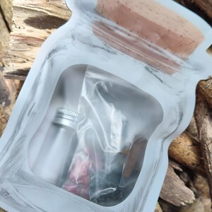 May include: A clear plastic pouch with a brown cork lid. Inside the pouch are a small silver bottle and a clear plastic bag with dried herbs and flowers.