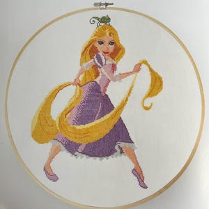 May include: Cross-stitch embroidery of a princess with long blonde hair, wearing a purple dress and pink top. The embroidery is set in a wooden hoop. The princess is holding her long hair, with a green chameleon on her head.