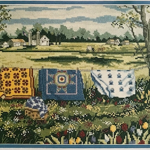 Peut inclure: Une broderie au point de croix représentant une scène rurale avec une ferme, un chariot tiré par des chevaux et trois courtepointes colorées accrochées à une corde à linge dans un champ de fleurs sauvages.