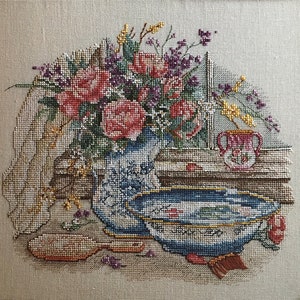 Peut inclure: Broderie au point de croix représentant une nature morte avec un arrangement floral dans un vase bleu et blanc. Un bol, une brosse et un petit pichet sont également visibles. Les fleurs sont roses, violettes et jaunes. Le fond est beige neutre.