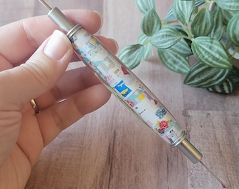 May include: A sewing awl with a decorative handle. The handle is clear with a colorful pattern of various images. The awl has a silver metal tip on each end. The awl is held in a hand.