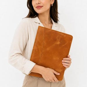 May include: A tan leather document portfolio featuring a smooth, matte finish and a vertical stitched seam detail along the front panel. The rectangular accessory is constructed from a single piece of leather with visible natural grain texture.
