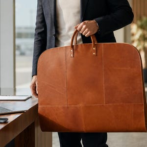 Puede incluir: Una funda para ropa grande y rectangular confeccionada en cuero liso de color tostado. El bolso cuenta con dos asas de cuero resistentes sujetas con remaches metálicos en la parte superior, una estructura rígida y costuras visibles.