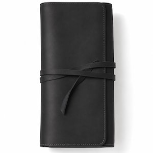 Puede incluir: Un estuche enrollable rectangular de cuero negro con un acabado mate liso y esquinas redondeadas. El estuche se asegura mediante una tira larga y fina de cuero negro que se envuelve horizontalmente alrededor del centro y se ata con un nudo.