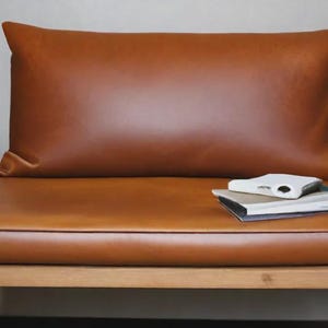 Peut inclure: Une chaise en bois dotée d'un dossier rectangulaire lisse et d'un coussin d'assise plat, tous deux recouverts d'un cuir marron chaud uni. Une petite pile de deux livres repose sur le côté droit du coussin d'assise, ajoutant un détail fonctionnel.