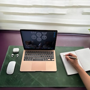 May include: A green leather desk mat with a laptop, notebook, pen, and wireless earbuds. The laptop screen shows a design of seven chakras. The desk mat has a small logo in the bottom center.