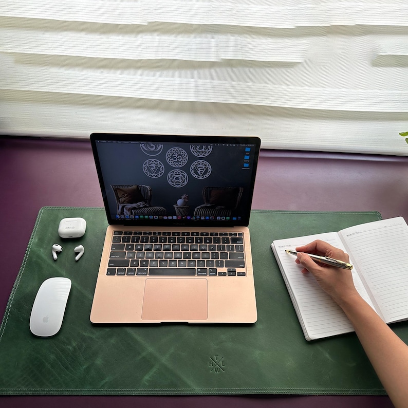 May include: A green leather desk mat with a laptop, notebook, pen, and wireless earbuds. The laptop screen shows a design of seven chakras. The desk mat has a small logo in the bottom center.