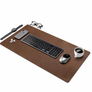 May include: A rectangular brown leather desk mat featuring a dark grey wireless keyboard, a silver computer mouse, a smartphone, a smartwatch, white wireless earbuds, black-rimmed spectacles, and a white ceramic mug filled with black coffee.