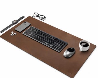 Può includere: Un sottomano rettangolare in pelle marrone con una tastiera wireless grigio scuro, un mouse per computer argentato, uno smartphone, uno smartwatch, auricolari wireless bianchi, occhiali con montatura nera e una tazza bianca con caffè nero.