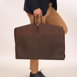 May include: A two-tone brown leather garment bag with a top handle and a buckle closure. The bag is rectangular and has a dark brown bottom section and a lighter brown top section. The bag is being held by a person.