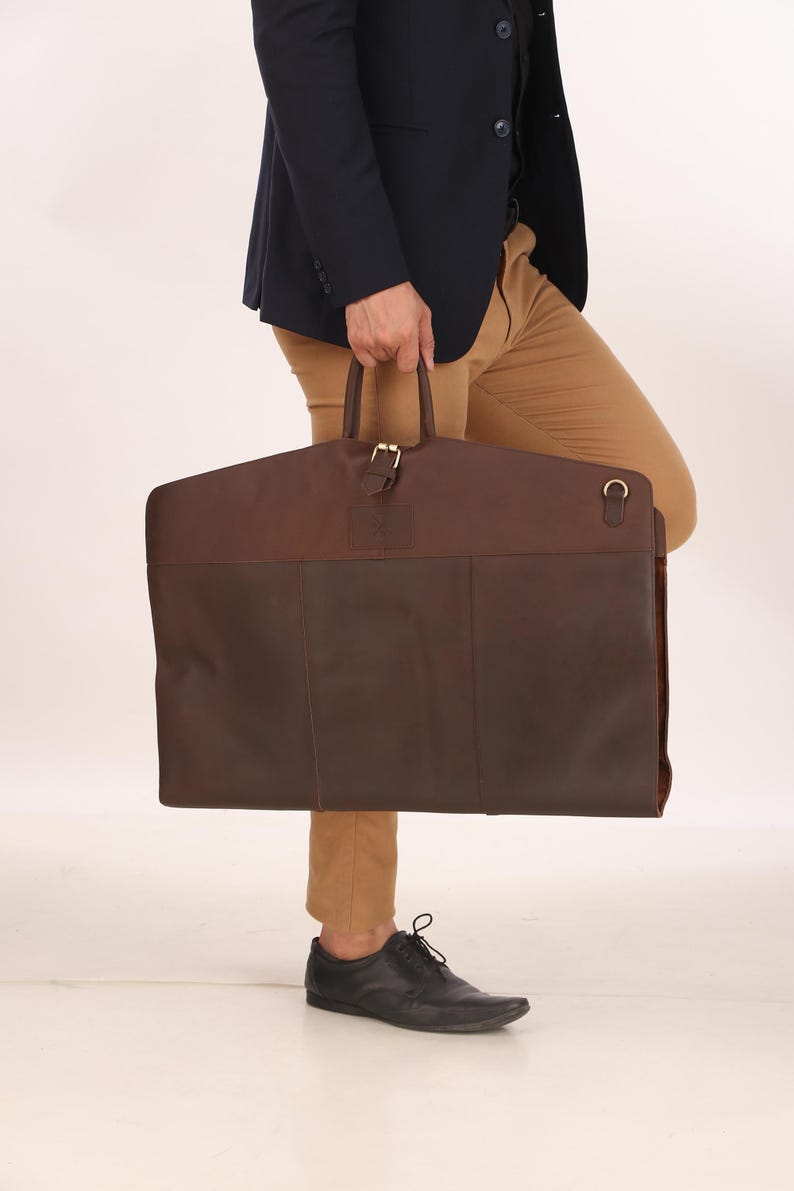 May include: A two-tone brown leather garment bag with a top handle and a buckle closure. The bag is rectangular and has a dark brown bottom section and a lighter brown top section. The bag is being held by a person.