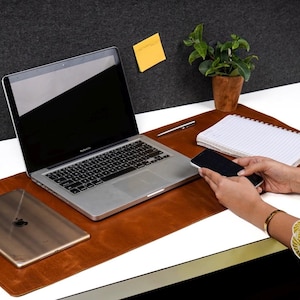 May include: A brown leather desk mat with a laptop, a tablet, a notepad, and a pen. The mat has a built-in phone holder and a slot for a pen.