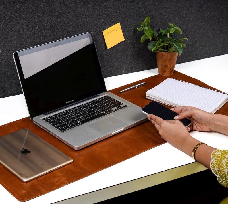 May include: A brown leather desk mat with a laptop, a tablet, a notepad, and a pen. The mat has a built-in phone holder and a slot for a pen.