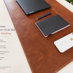 May include: A rectangular brown leather desk mat with visible edge stitching. The mat features the text Premium Leather Desk Mat Edge Stitching, along with icons and text describing edge stitching, desk protection, and handcrafted construction.