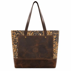 Peut inclure: Un sac cabas doté d'une base en cuir marron foncé et d'une poche centrale avec un logo circulaire en relief. Les côtés et le panneau supérieur présentent un motif floral détaillé dans des tons noir, doré et crème. Il possède deux longues anses.