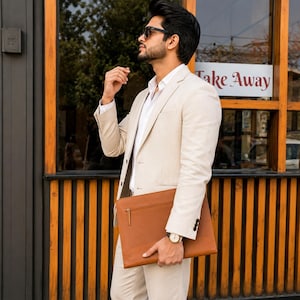 May include: A beige blazer worn over a white collared shirt, paired with matching beige trousers. A tan leather document portfolio with a gold zip is held under the arm. The ensemble includes dark sunglasses and a wristwatch with a white face.