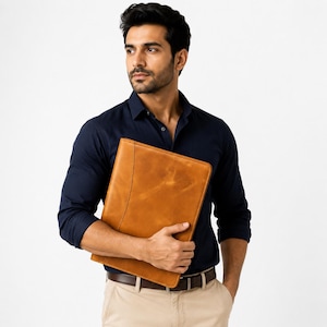 May include: A tan leather document portfolio with a smooth, matte finish and visible stitching along the side, held against a dark navy blue long-sleeved button-down shirt and light khaki trousers with a dark brown leather belt.