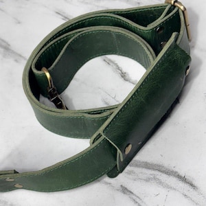 May include: A forest green leather bag strap featuring a sliding shoulder pad, antique brass-toned metal hardware, adjustable buckle, and swivel lobster clasps. The strap is constructed with visible stitching and secured with small metal rivets.