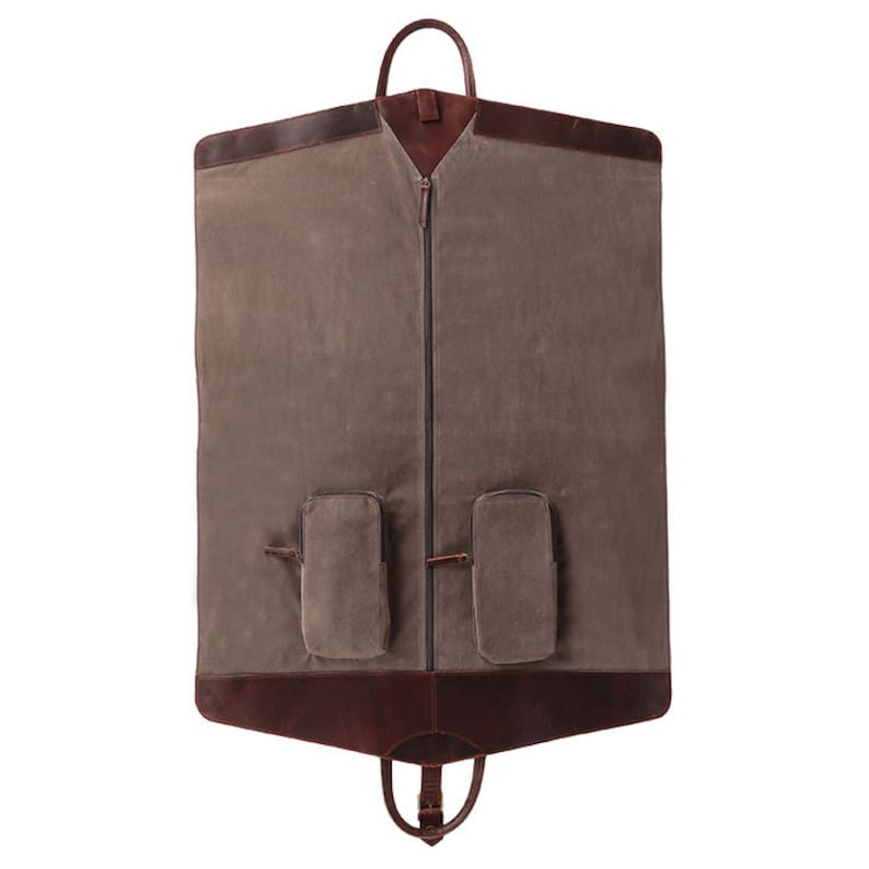 May include: A brown canvas garment bag with leather accents. The bag features a full-length zipper, two exterior pockets, and a top handle. The bottom has a handle for carrying. The bag is designed to protect clothing during travel.