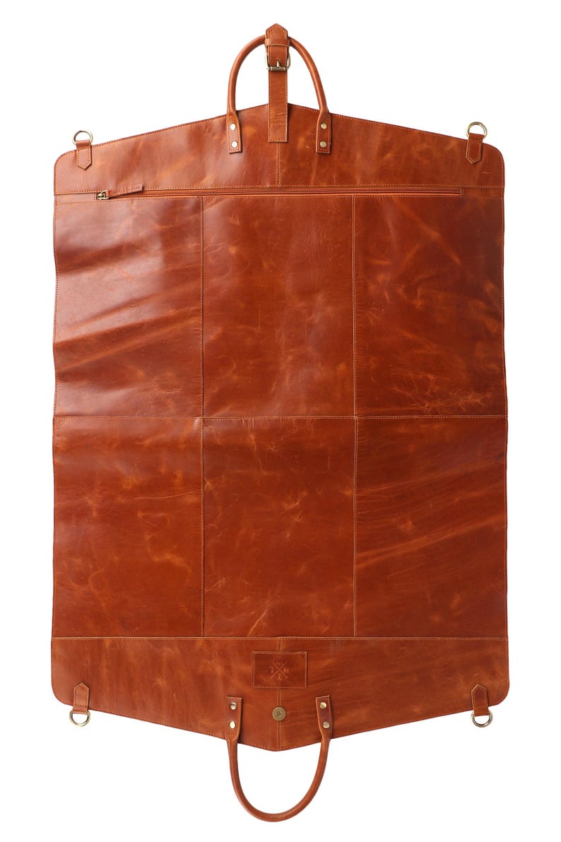 May include: A brown leather garment bag with a top handle and a bottom handle. The bag has a zipper closure and several metal D-rings. The leather has a distressed appearance. The bag is designed to carry clothing.