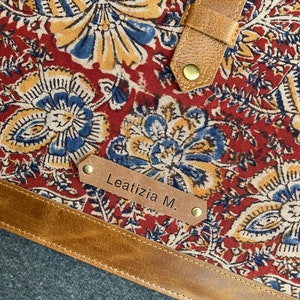 May include: Close-up of a tote bag with a floral pattern in red, blue, and yellow. The bag has brown leather straps and trim, with the name "Leatizia M." on a leather tag.