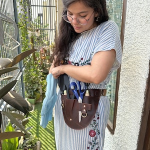 Puede incluir: Un cintur&oacute;n de herramientas de jardiner&iacute;a de cuero marr&oacute;n oscuro con m&uacute;ltiples bolsillos y trabillas, que sostiene unas tijeras de mango azul y una peque&ntilde;a pala negra. El cintur&oacute;n se lleva sobre un vestido de rayas blancas y azules bordado.