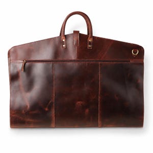 Dark brown leather garment bag with rounded top handles, front zip pocket, stitched leather panels and a brass-tone side D-ring on a clean white background.
