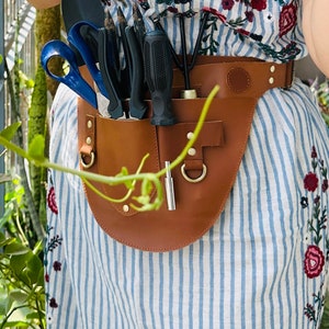 Puede incluir: Un cintur&oacute;n utilitario de cuero color canela con m&uacute;ltiples bolsillos y herrajes en tono lat&oacute;n, usado sobre un vestido a rayas blancas y azules con bordados florales. El cintur&oacute;n sostiene varias herramientas de jardiner&iacute;a.