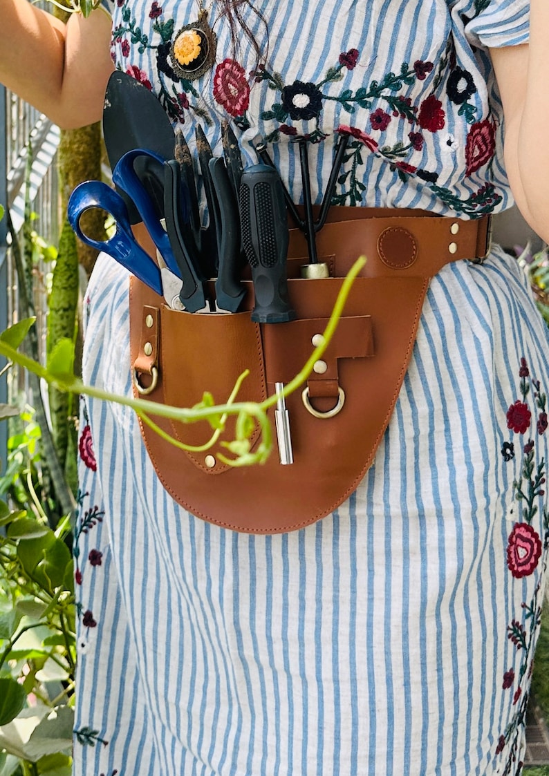 Puede incluir: Un cintur&oacute;n utilitario de cuero color canela con m&uacute;ltiples bolsillos y herrajes en tono lat&oacute;n, usado sobre un vestido a rayas blancas y azules con bordados florales. El cintur&oacute;n sostiene varias herramientas de jardiner&iacute;a.